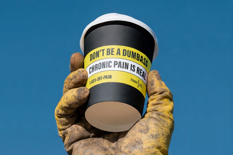 Coffee cup that reads "Don't be a Dumbass chronic pain is real" held in the gloved hand of a tradesperson.